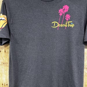 Könnte beinhalten: Dunkelgraues T-Shirt mit einer Grafik von rosa Palmen und dem Schriftzug "Desert Trip" in gelber Schrift. Der linke Ärmel hat ein farbenfrohes Zungenlogo. Das Shirt hat einen Rundhalsausschnitt und kurze Ärmel.