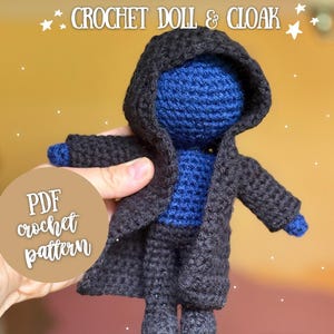 May include: A handmade crochet doll wearing a black cloak with a hood. The doll's head and chest are blue, and the cloak is dark gray. The image includes text that says "CROCHET DOLL & CLOAK" and "PDF crochet pattern".