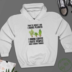 May include: White hoodie with a black graphic print that says "That's what I do I have plants I drink coffee I hate people and I know things". The graphic also includes three potted plants.