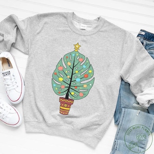 May include: A gray sweatshirt with a green monstera leaf shaped Christmas tree with colorful ornaments. The tree is in a red and yellow pot. The text "Just More Plants" is on a white sticker on the bottom right corner of the image.