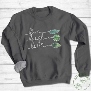 May include: A dark grey sweatshirt with the words "live laugh love" written in white cursive. The words are separated by three green leaves.
