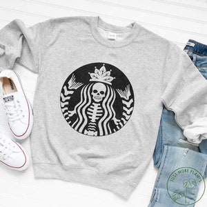May include: A grey sweatshirt featuring a black and white graphic of a skeleton wearing a crown and holding a cup. The graphic is a parody of the Starbucks logo.