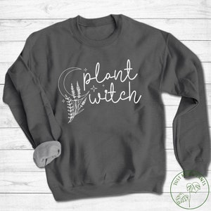 May include: A dark grey sweatshirt with the text "plant witch" in white lettering. The text is accompanied by a crescent moon and stylised floral design.