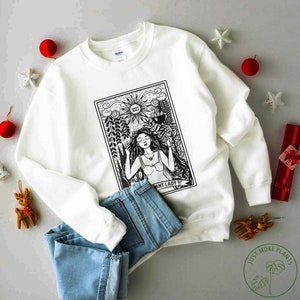 May include: White sweatshirt featuring a black and white illustration of a tarot card. The card is the High Priestess and depicts a woman with long hair and a crown. The text on the card reads "High Priestess".