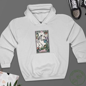 May include: White hoodie featuring a graphic of a skeleton watering plants. The graphic is a tarot card with the text "The Plant Lady".