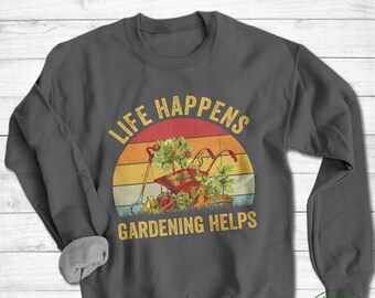 May include: A dark gray sweatshirt with a vintage-style graphic that says "Life Happens Gardening Helps". The graphic features a wheelbarrow with plants and gardening tools in front of a sunset.