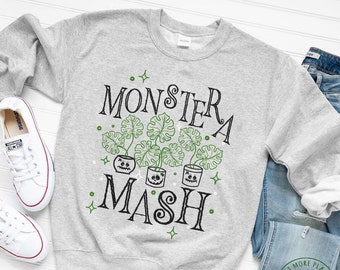 May include: A gray sweatshirt with the text "Monstera Mash" in black lettering. The text is surrounded by green monstera leaves with jack-o-lantern faces. There are small white stars scattered around the text.