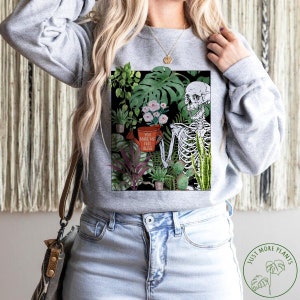 May include: A grey sweatshirt featuring a graphic print of a skeleton holding a watering can with the text "You make me feel alive". The skeleton is surrounded by various plants. The sweatshirt has a crew neck and long sleeves.