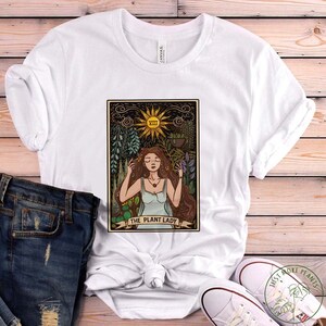 Pode incluir: Camiseta branca com uma ilustra&ccedil;&atilde;o de carta de tar&ocirc; vintage de uma mulher cercada por plantas. O cart&atilde;o diz "The Plant Lady".