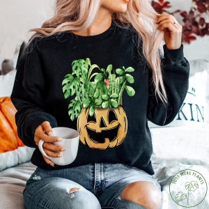 May include: Black sweatshirt featuring a graphic of a jack-o'-lantern with a variety of green plants growing out of the top. The text "Just More Plants" is on a green circle with a leaf design.
