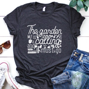 Może przedstawiać: Szary t-shirt z białym nadrukiem, na którym widnieje napis "The garden is calling and I must go". Nadruk jest otoczony małymi białymi ilustracjami narzędzi ogrodniczych i roślin.