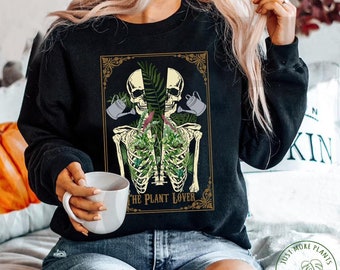 May include: A black sweatshirt with a graphic of two skeletons with plants growing out of their ribcages. The graphic is surrounded by a gold frame and the text "The Plant Lover" is below the skeletons. The sweatshirt is being worn by a person with long blonde hair.