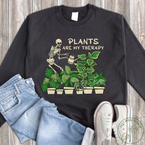 May include: A black sweatshirt featuring a skeleton watering plants. The text "Plants are my therapy" is printed on the sweatshirt.