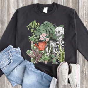 May include: Black sweatshirt featuring a skeleton holding a watering can and surrounded by plants. The text on the sweatshirt reads "You make me feel alive".
