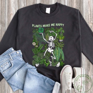 Może przedstawiać: Czarna bluza z nadrukiem szkieletu tańczącego wśród zielonych roślin. Na przodzie bluzy widnieje napis "PLANTS MAKE ME HAPPY".
