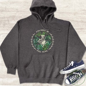May include: A dark grey hoodie featuring a graphic of a skeleton watering plants. The graphic is enclosed in a circle with the text "You make me feel alive".