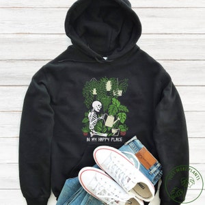 May include: Black hoodie featuring a graphic of a skeleton watering plants. The graphic reads "In my happy place".