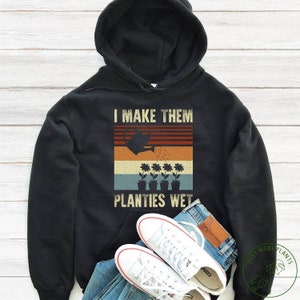 May include: Black hoodie featuring a vintage-style graphic that reads "I make them planties wet". The graphic depicts a watering can pouring water onto four potted plants with flowers.