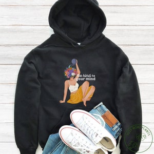 May include: Black hoodie with an illustration of a person with a floral head, holding a blue object, and the text "Be kind to your mind." The person is wearing a white top and yellow skirt. White trainers and blue jeans are also visible.