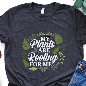 May include: A dark grey t-shirt with a white graphic that says "My Plants Are Rooting For Me". The graphic is surrounded by green leaves and flowers.