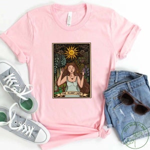 Pode incluir: Camiseta rosa com um design de carta de tar&ocirc; vintage que apresenta uma mulher cercada por plantas e o texto "The Plant Lady".