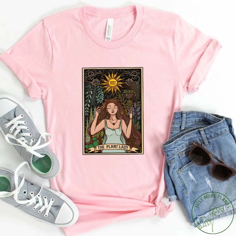 Pode incluir: Camiseta rosa com um design de carta de tar&ocirc; vintage que apresenta uma mulher cercada por plantas e o texto "The Plant Lady".