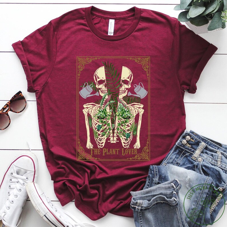Peut inclure: T-shirt bordeaux avec un graphisme unique. Le motif pr&eacute;sente un squelette avec des plantes dans la cage thoracique, deux cr&acirc;nes et des arrosoirs. Le texte "THE PLANT LOVER" est affich&eacute; en dessous. Des lunettes de soleil et des baskets sont visibles.