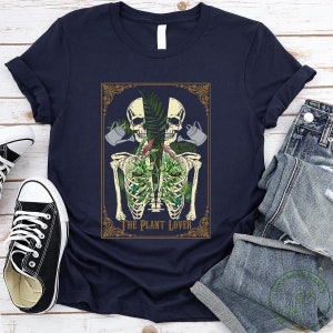 Peut inclure: T-shirt bleu marine avec un motif de squelette avec des plantes dans la cage thoracique, deux cr&acirc;nes et des arrosoirs. Le texte "THE PLANT LOVER" est en dessous. Le t-shirt est pli&eacute;, avec un jean et des baskets en arri&egrave;re-plan.