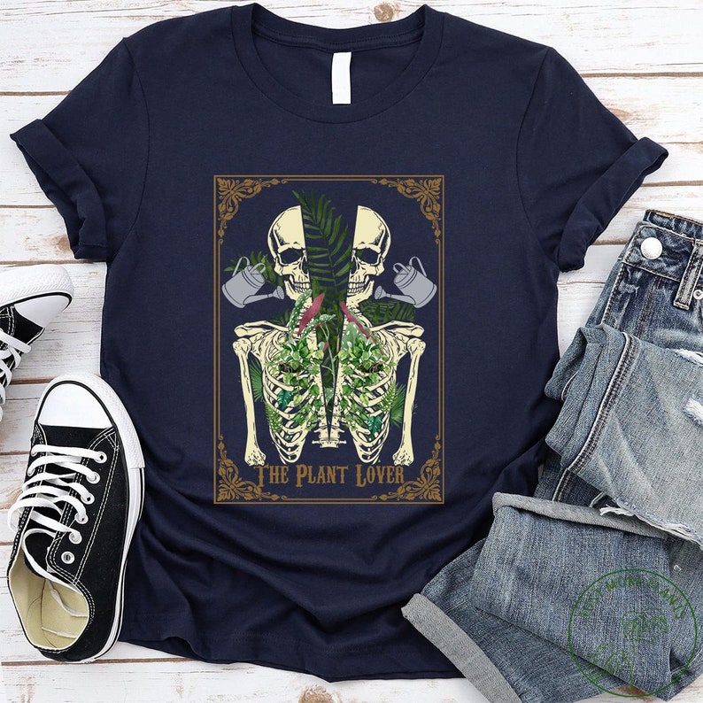 Peut inclure: T-shirt bleu marine avec un motif de squelette avec des plantes dans la cage thoracique, deux cr&acirc;nes et des arrosoirs. Le texte "THE PLANT LOVER" est en dessous. Le t-shirt est pli&eacute;, avec un jean et des baskets en arri&egrave;re-plan.