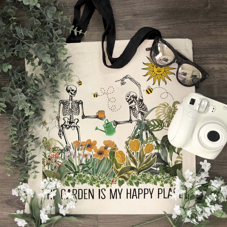 May include: A white tote bag with a black handle and a design of two skeletons gardening. The skeletons are holding a watering can and a cup. The text on the bag reads "My Garden is My Happy Place".