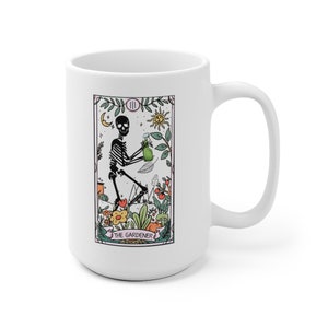 May include: White ceramic mug with a black and white illustration of a skeleton holding a watering can. The illustration is surrounded by a pink border and the text "The Gardener" is below the illustration.