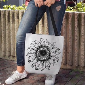 Peut inclure: Un sac fourre-tout blanc avec un dessin au trait noir et blanc d'un tournesol. Le sac a des bretelles noires et est port&eacute; par une personne portant un jean bleu.