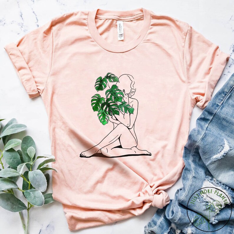 May include: A light pink t-shirt featuring a graphic of a woman sitting with a large green monstera deliciosa plant in front of her face.