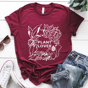 May include: A maroon t-shirt with a white graphic design of plants and a squirrel. The text "PLANT LOVER" is featured on the shirt.