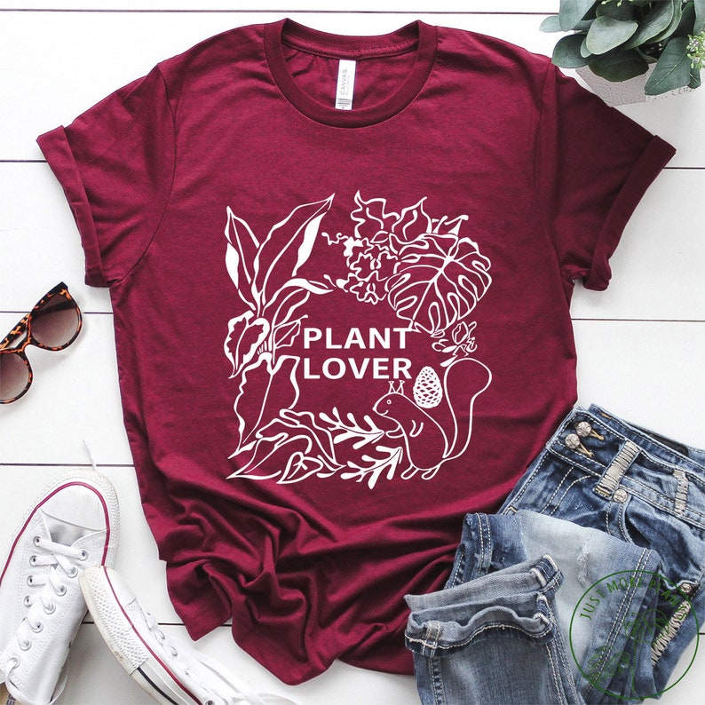 May include: A maroon t-shirt with a white graphic design of plants and a squirrel. The text "PLANT LOVER" is featured on the shirt.