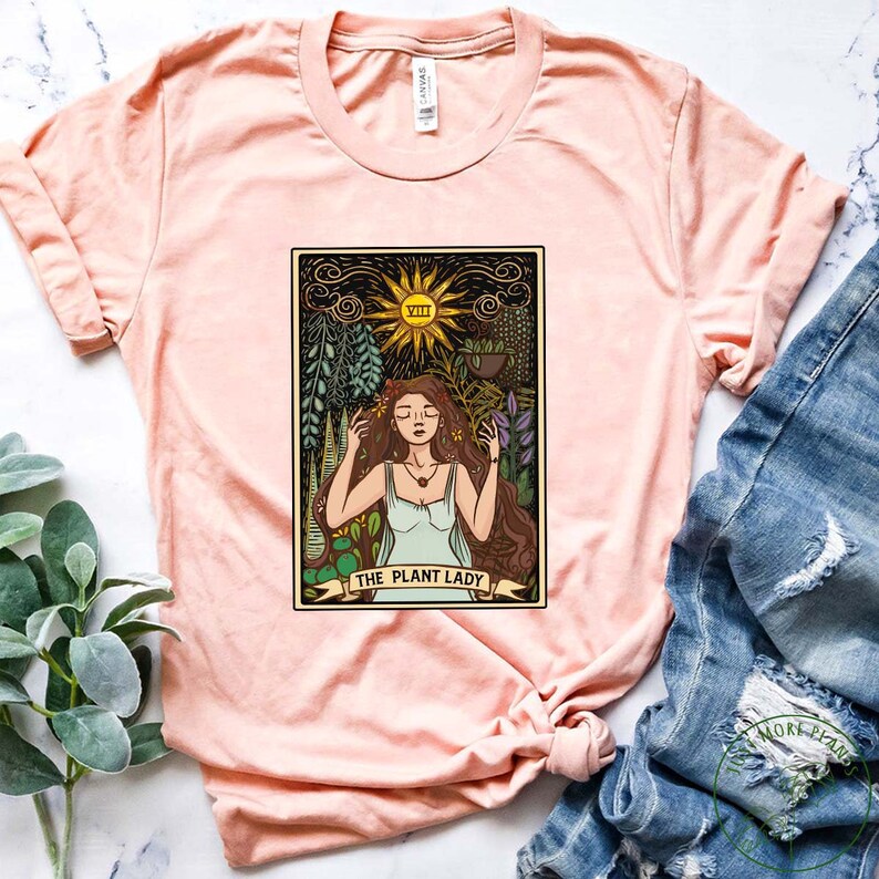 Pode incluir: Uma camiseta cor de p&ecirc;ssego com um design de carta de tar&ocirc; vintage que apresenta uma mulher cercada por plantas e o texto "The Plant Lady".