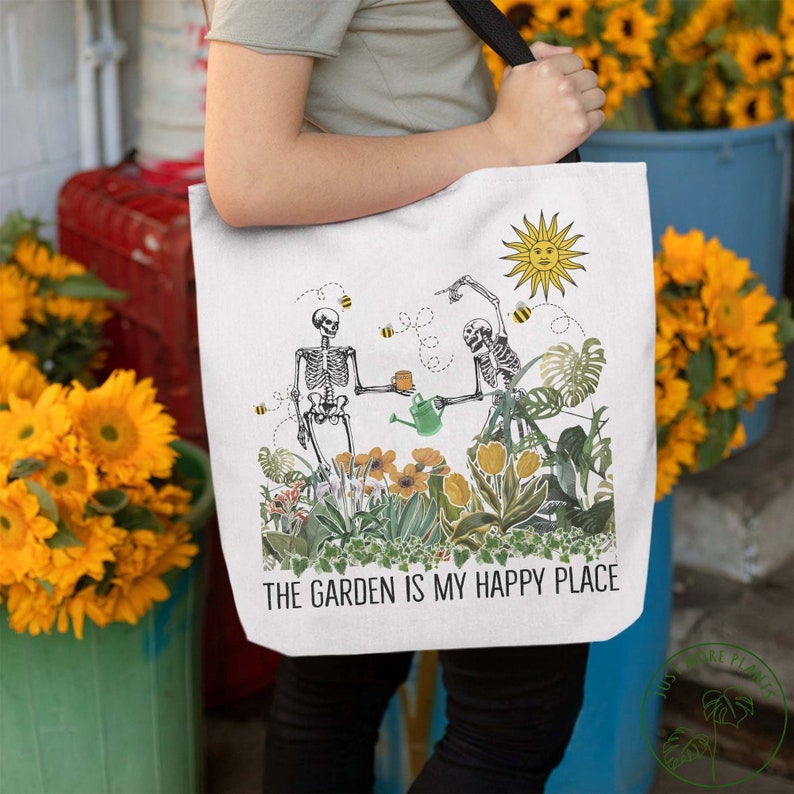 May include: White tote bag with a black handle. The bag features a graphic of two skeletons gardening with a watering can and a cup of tea. The text "The Garden is My Happy Place" is printed on the bag.
