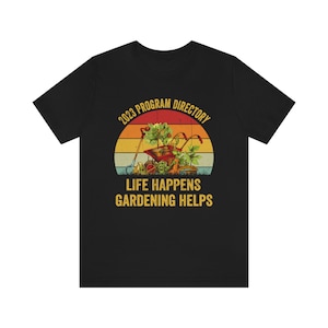May include: Black t-shirt with a vintage-style graphic of a wheelbarrow full of gardening tools and plants. The graphic is set against a sunset background with the text "2023 Program Directory" above and "Life Happens Gardening Helps" below.