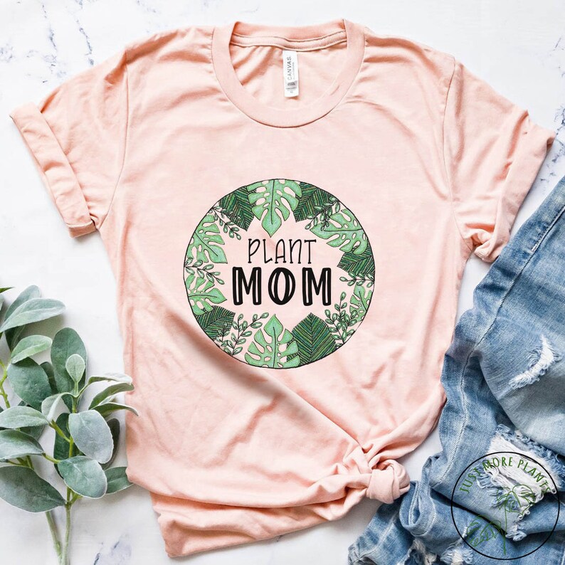 May include: A light pink t-shirt with a green circle design featuring the text "PLANT MOM" surrounded by leaves.