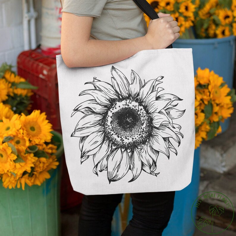 Peut inclure: Un sac fourre-tout blanc avec un dessin au trait noir et blanc d'un tournesol. Le sac a une sangle noire et est tenu par une personne.