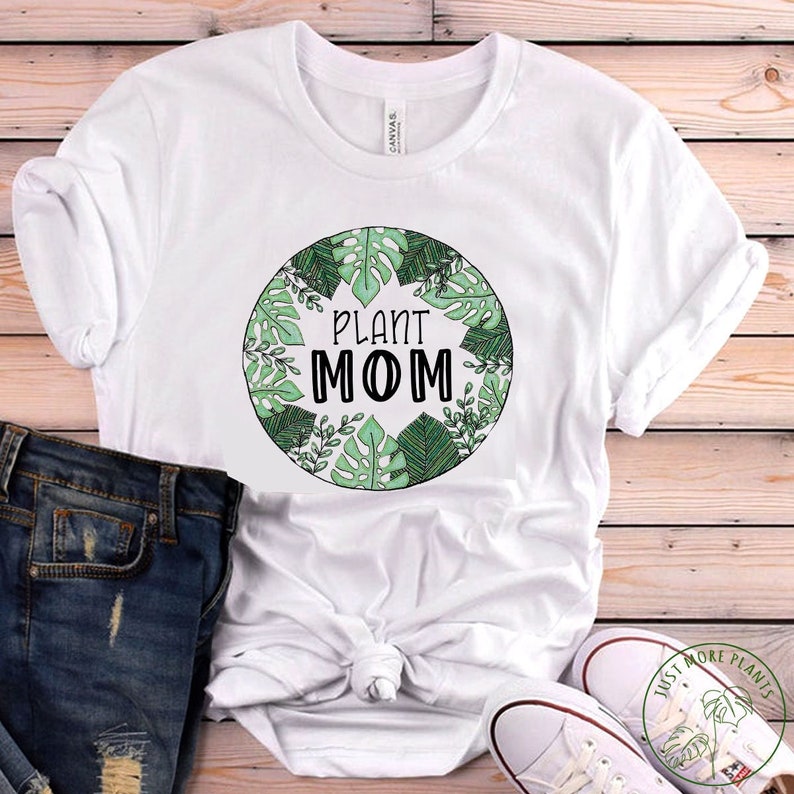 May include: White t-shirt with a green circle graphic that says "Plant Mom" surrounded by green leaves. The shirt is laying on a wooden surface with a pair of white sneakers and blue jeans.