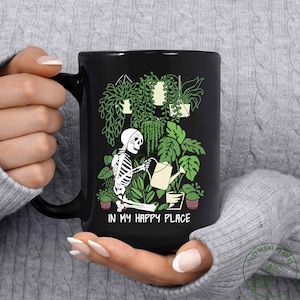 May include: Black coffee mug with a skeleton watering plants. The mug has the text "In my happy place" on the bottom.