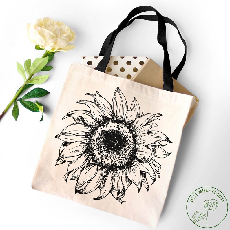 Peut inclure: Un sac fourre-tout en toile blanche avec des sangles noires pr&eacute;sente un dessin au trait noir et blanc d'un tournesol. Le texte "Just More Plants" est imprim&eacute; en vert sur un cercle blanc dans le coin inf&eacute;rieur droit du sac.
