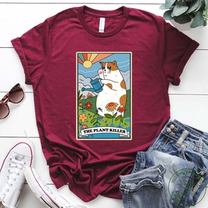 May include: A maroon t-shirt with a cartoon illustration of a cat holding a watering can in a garden setting. The text "The Plant Killer" is printed below the image.