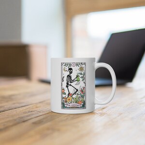 May include: White ceramic mug with a colorful illustration of a skeleton holding a watering can and surrounded by flowers. The text "The Gardener" is printed below the image.