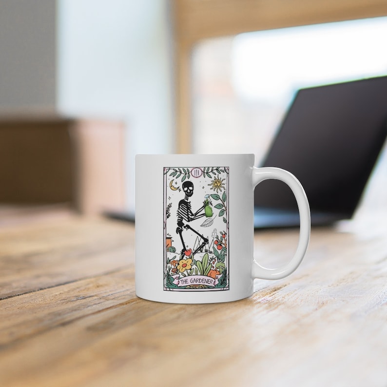 May include: White ceramic mug with a colorful illustration of a skeleton holding a watering can and surrounded by flowers. The text "The Gardener" is printed below the image.