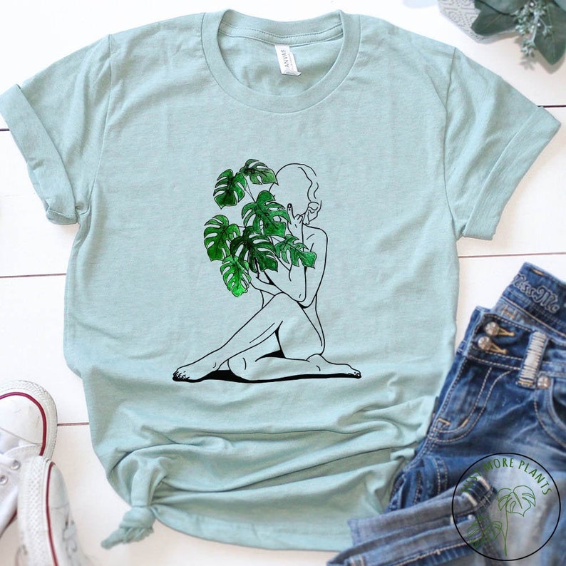 May include: A light blue t-shirt featuring a black line drawing of a person holding a green monstera plant. The plant is in the foreground and the person is in the background.