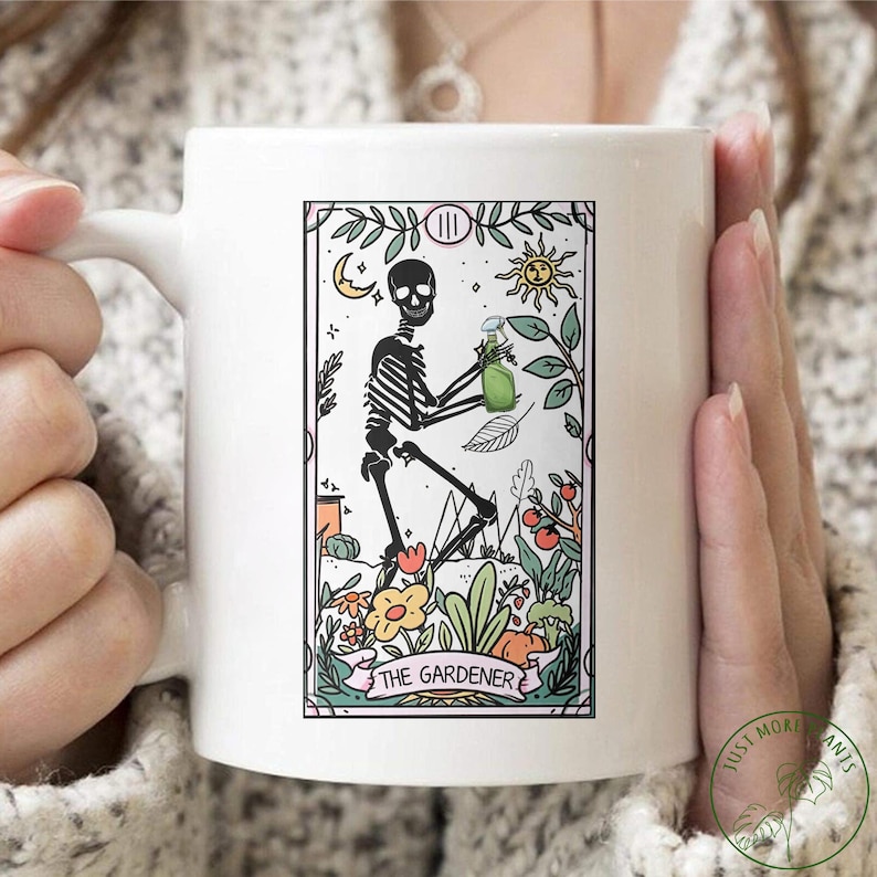 May include: White ceramic mug with a black and white illustration of a skeleton holding a spray bottle in a garden setting. The illustration is surrounded by a pink banner with the text "THE GARDENER".