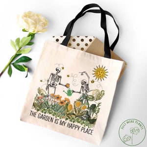 May include: A white tote bag with black handles features a design of two skeletons gardening. The skeletons are holding a watering can and a cup of tea. The text "The Garden is My Happy Place" is printed on the bag. A green circle with the text "Just More Plants" is in the bottom right corner.