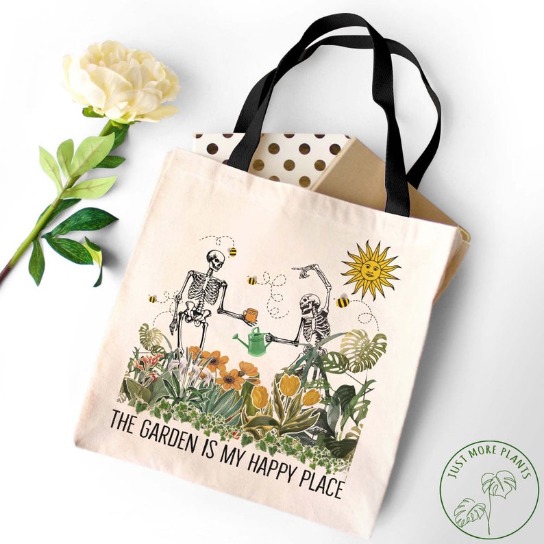 May include: A white tote bag with black handles features a design of two skeletons gardening. The skeletons are holding a watering can and a cup of tea. The text "The Garden is My Happy Place" is printed on the bag. A green circle with the text "Just More Plants" is in the bottom right corner.